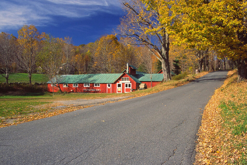 a picturesque red building with a green roof emulates the feel of an old farm building. there are trees with autumn foliage all around and a road passes by